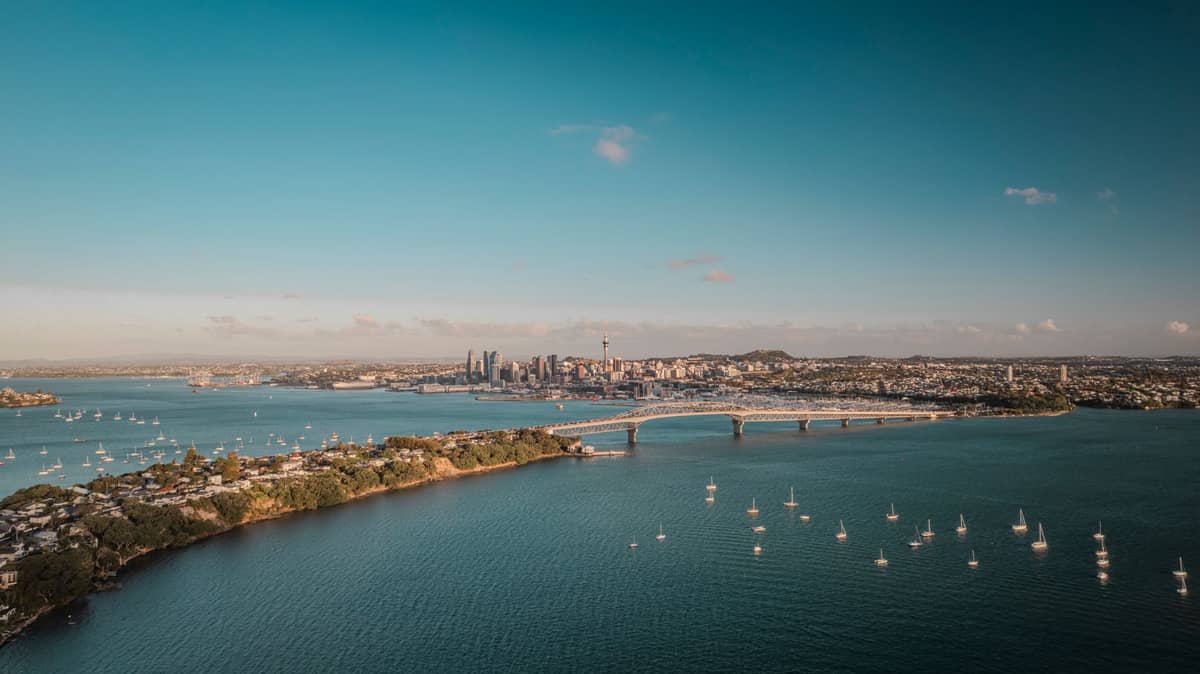 เส้นขอบฟ้าเมืองโอ๊คแลนด์เหนือน้ำท่าเรือสีฟ้า มีเรือลอยอยู่ประปราย Auckland Harbour Bridge และแนวชายฝั่งภายใต้ท้องฟ้าแจ่มใส