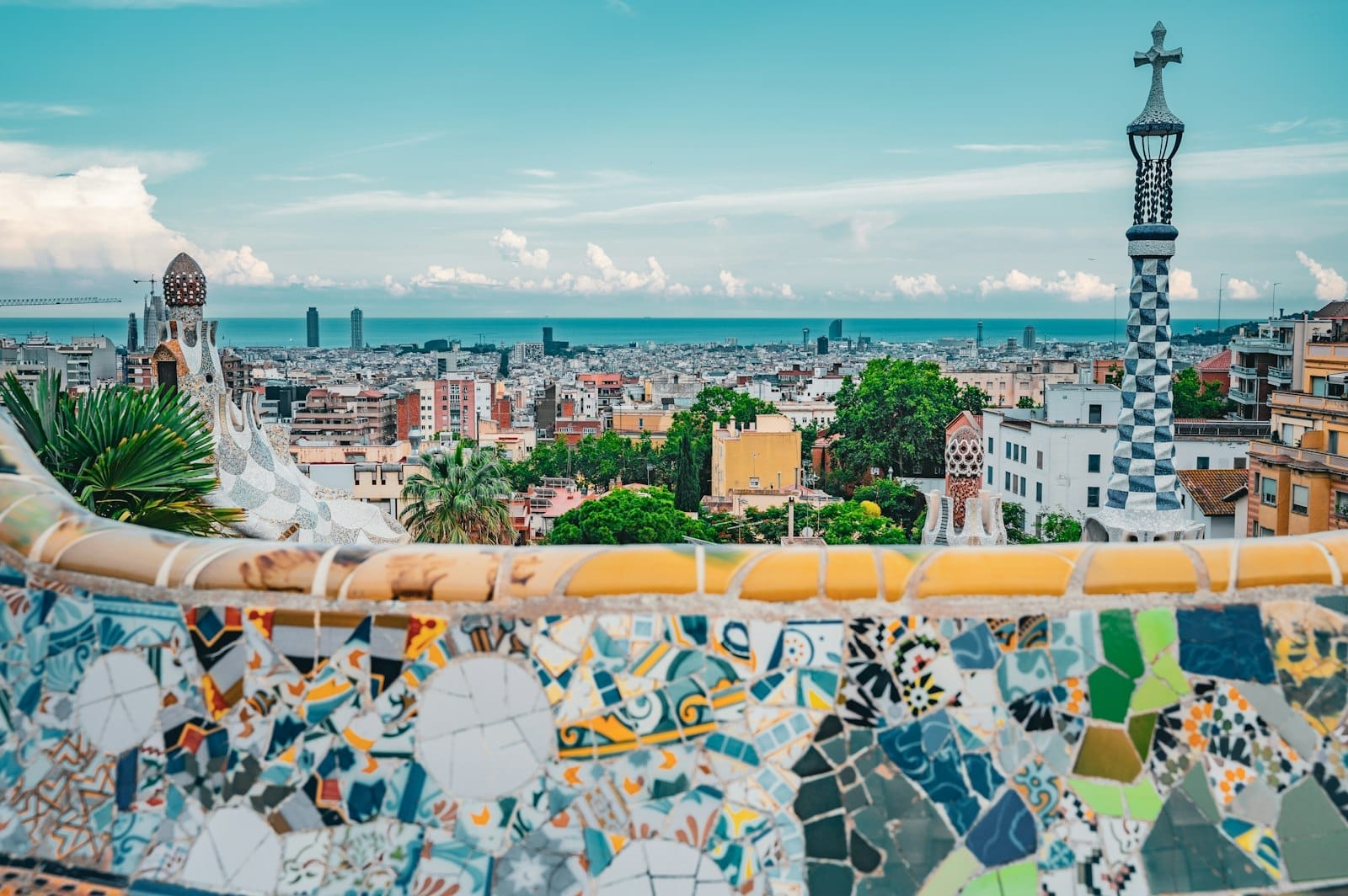 Colorful mosaic-tiled terrace in Park Güell overlooking the cityscape and Mediterranean Sea of Barcelona on a bright, clear day.