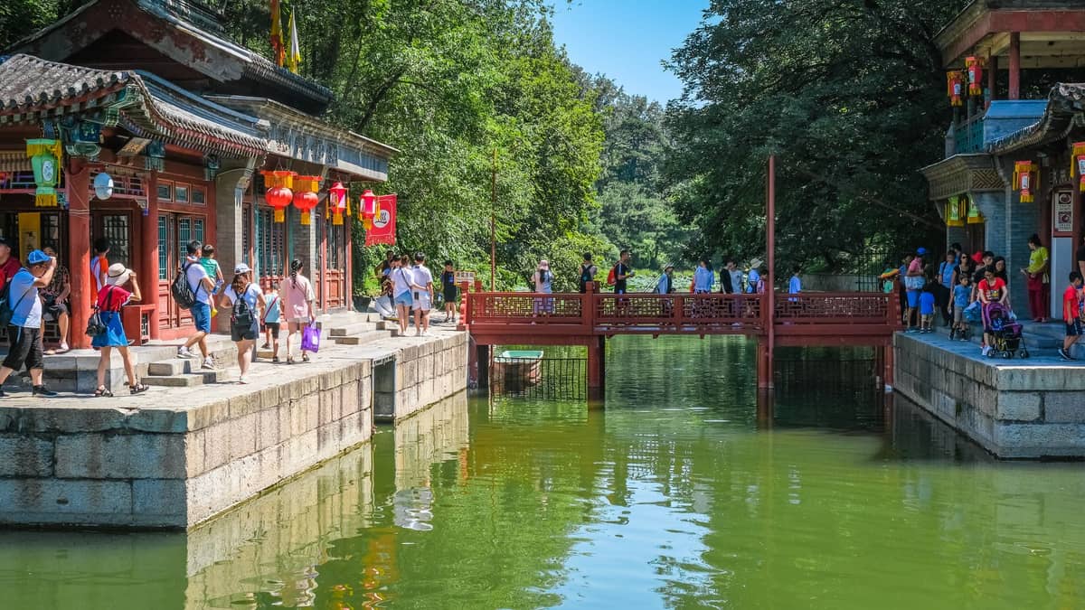 Des foules se promènent le long et sur un pavillon chinois traditionnel et un pont rouge au-dessus d'une eau verte sous un soleil d'été éclatant avec des arbres luxuriants à Pékin.