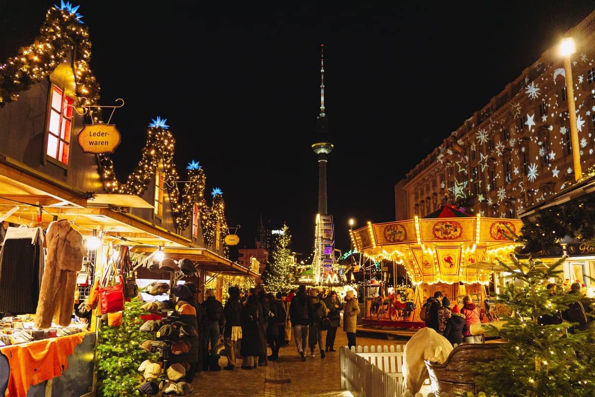 Berliner Weihnachtsmarkt bei Nacht mit festlicher Beleuchtung, dekorierten Ständen, einem Karussell und dem Fernsehturm im Hintergrund