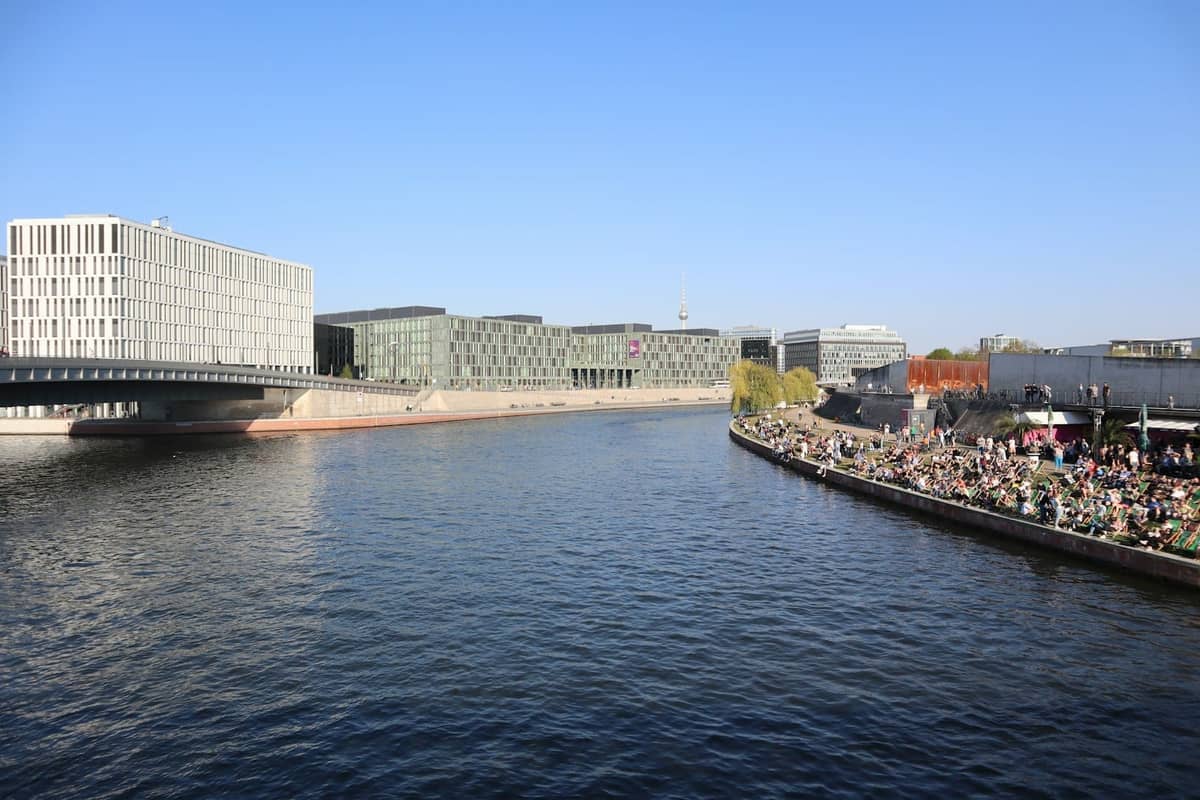 Blick auf ein belebtes Berliner Flussufer im Sommer mit entspannenden Menschenmengen an der Spree, modernen Gebäuden und klarem blauem Himmel.