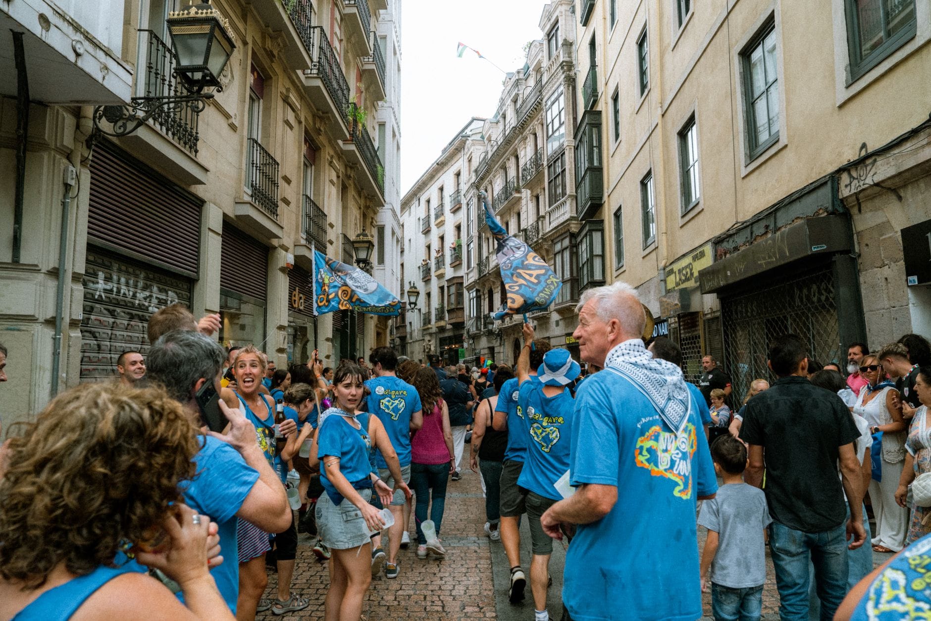 Uma animada cena de rua de verão em Bilbao, com pessoas em camisas azuis festivas, bandeiras e faixas criando uma atmosfera movimentada e comemorativa.