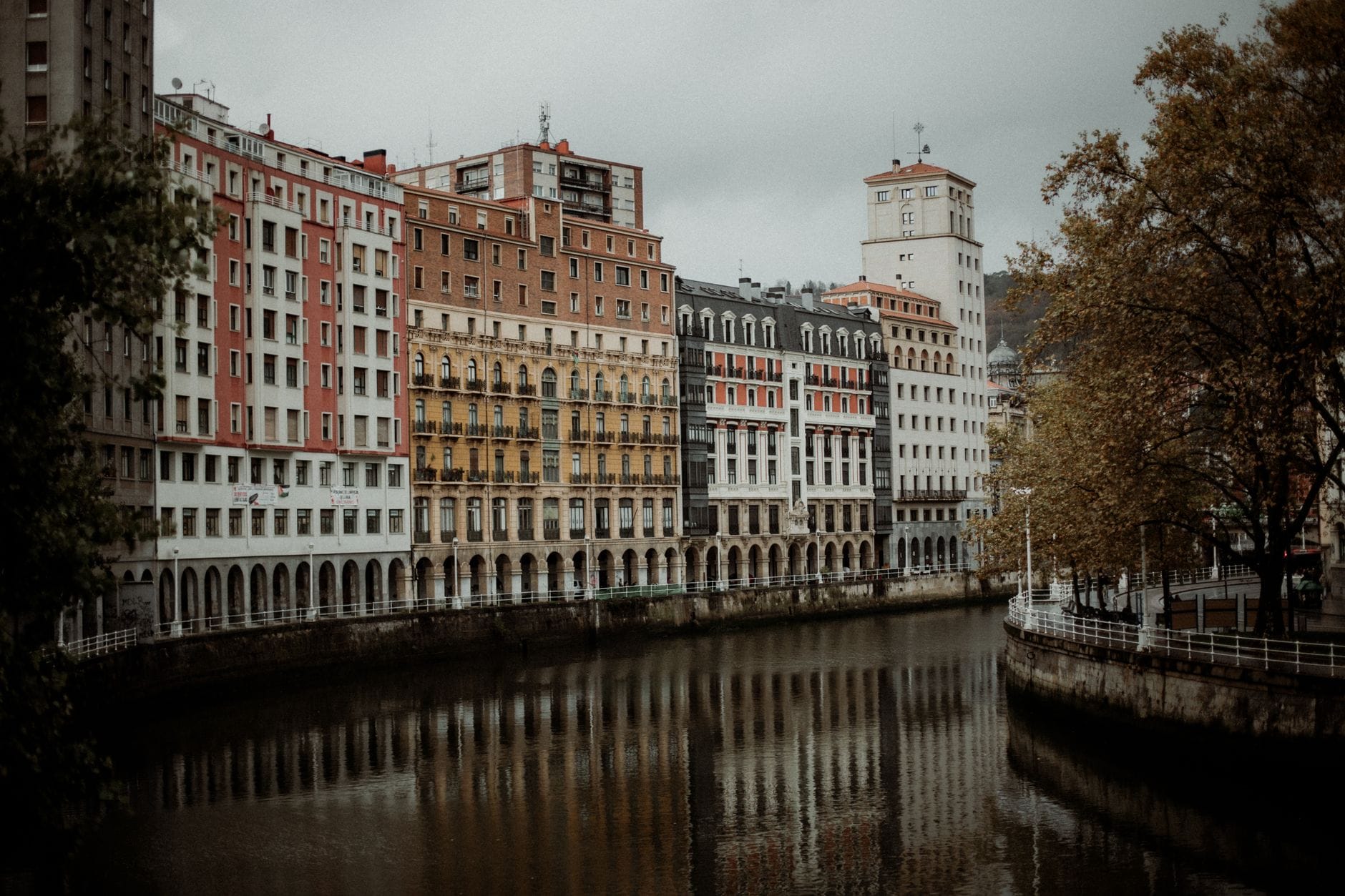 Cena outonal melancólica com edifícios à beira do rio em Bilbao, céu nublado e árvores com folhas murchas refletidas na água.