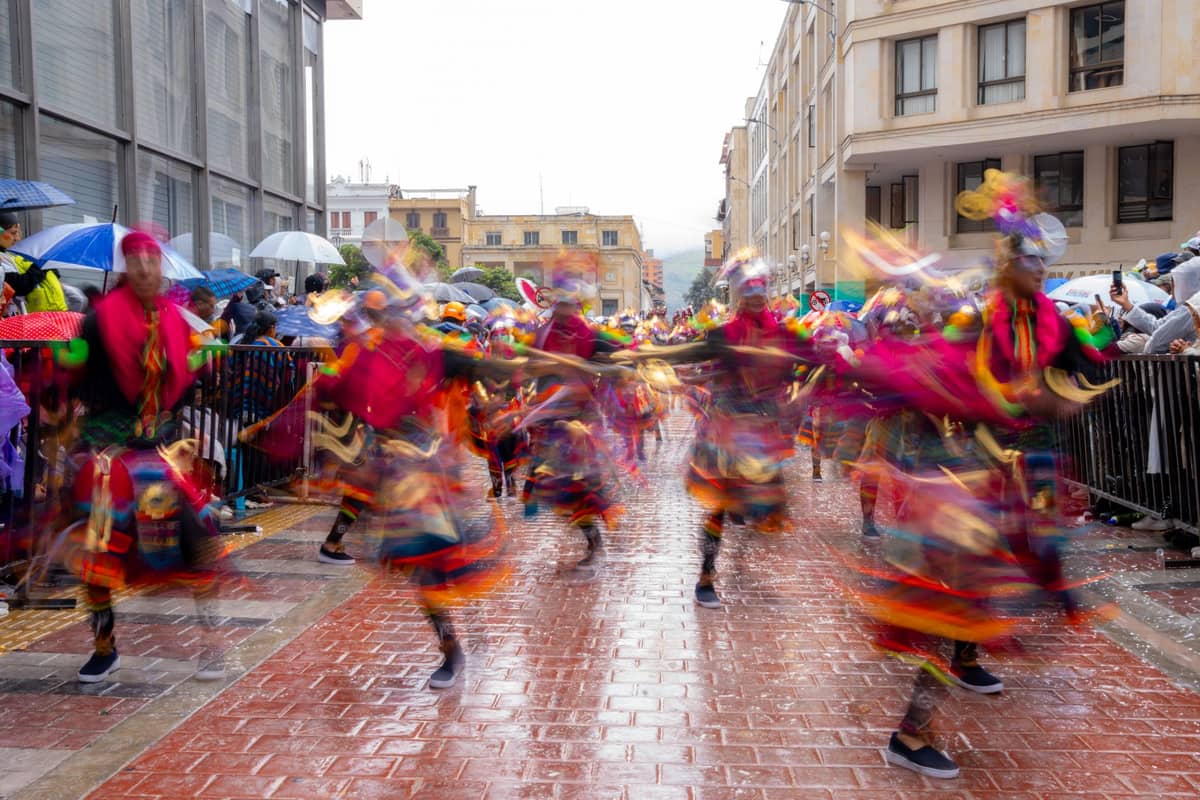 Colorful costumed dancers performing in a lively outdoor street festival in Bogotá as crowds watch, under umbrellas on a rainy day.