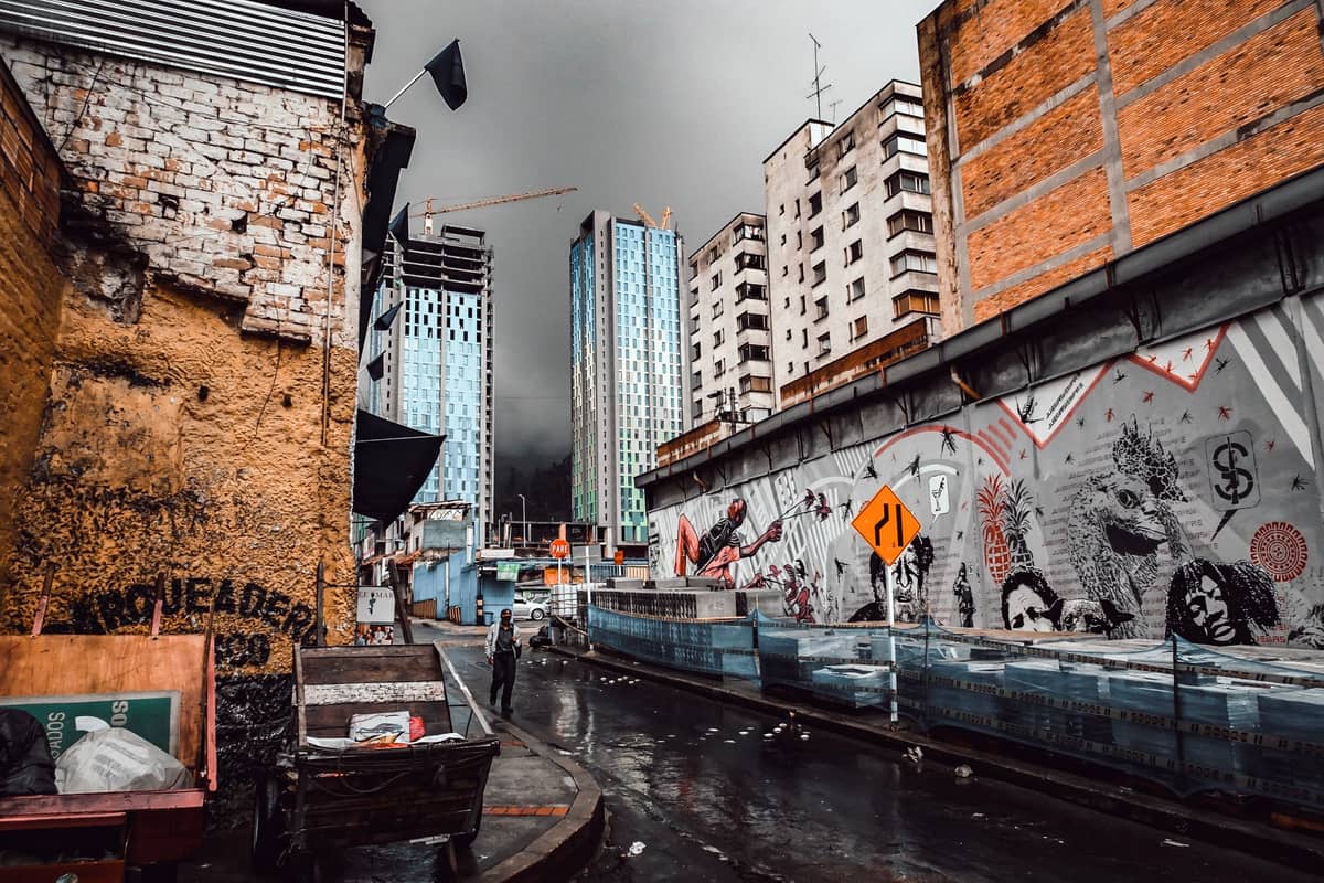 Rainy urban street in Bogotá with puddles, tall buildings, and wall murals under a cloudy sky.