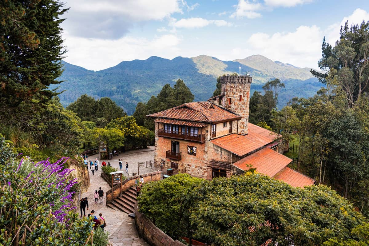 Sunny landscape view from Monserrate hill in Bogotá with clear mountains and people walking near a historic building.