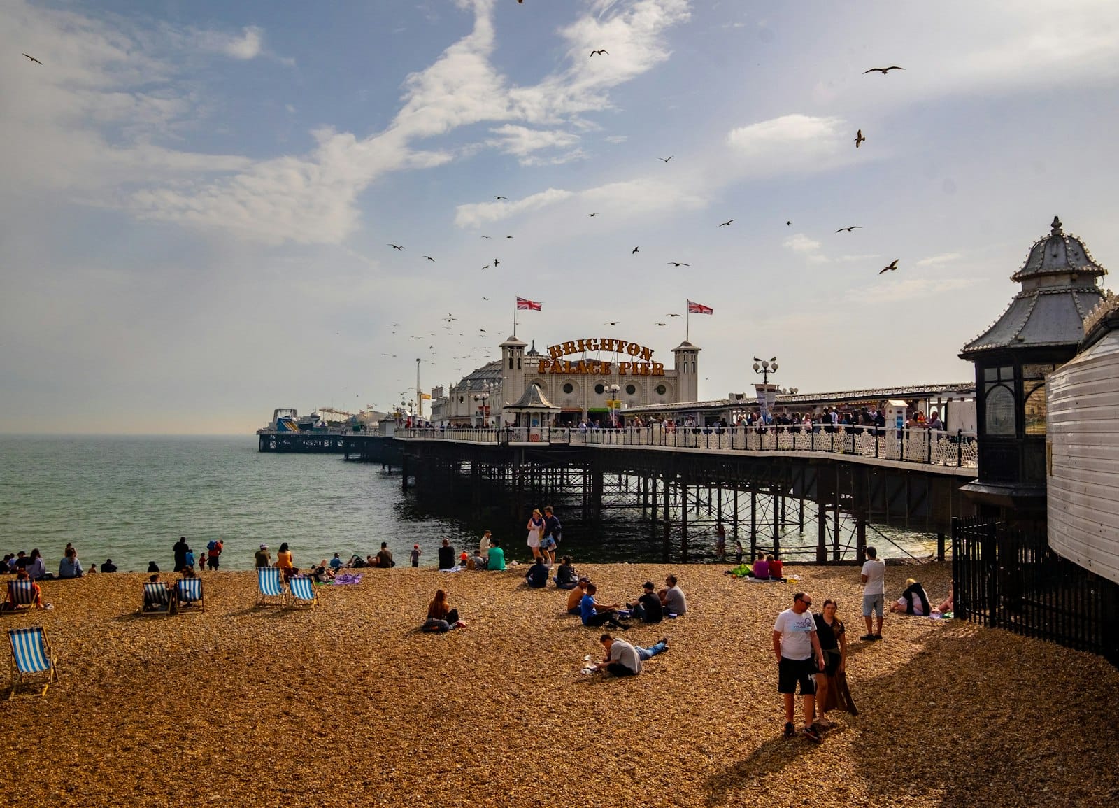 Brighton Pier viewed from the pebbly beach on a partly sunny day with people relaxing and enjoying the seaside atmosphere.