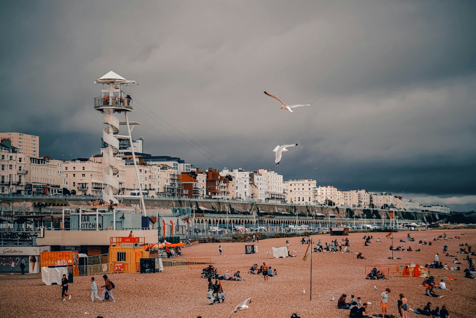 Cielos nublados sobre la playa de Brighton, con personas sentadas en los guijarros y gaviotas volando, creando una escena costera fresca y cubierta.