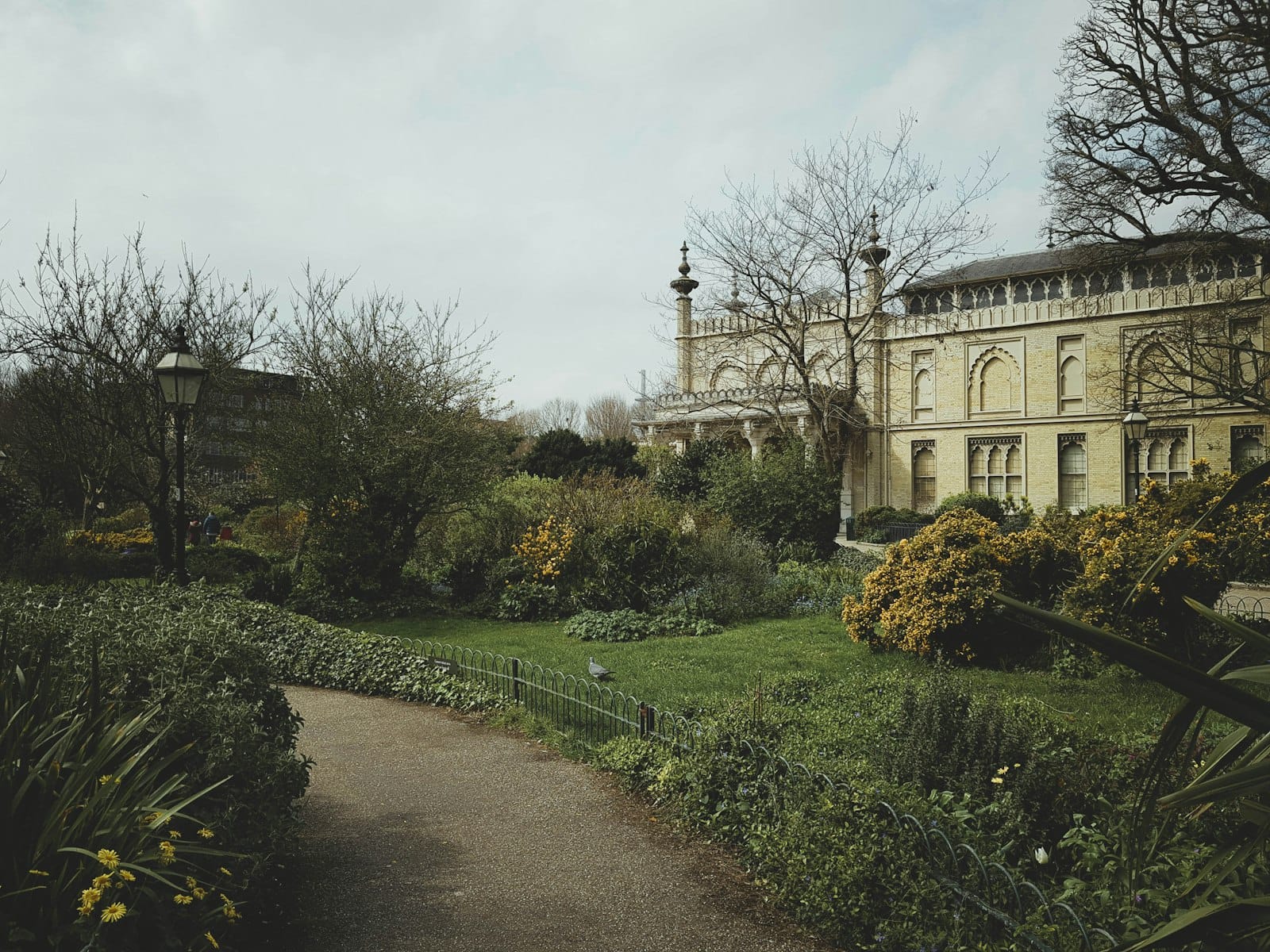Vista del Royal Pavilion con jardines primaverales en ciernes, arbustos con hojas y un cielo nublado a lo largo de un camino de Brighton.