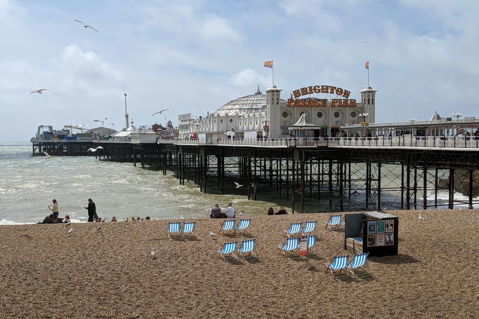 Vista amplia del Brighton Palace Pier con tumbonas en la playa de guijarros y personas disfrutando del paseo marítimo bajo cielos nublados.