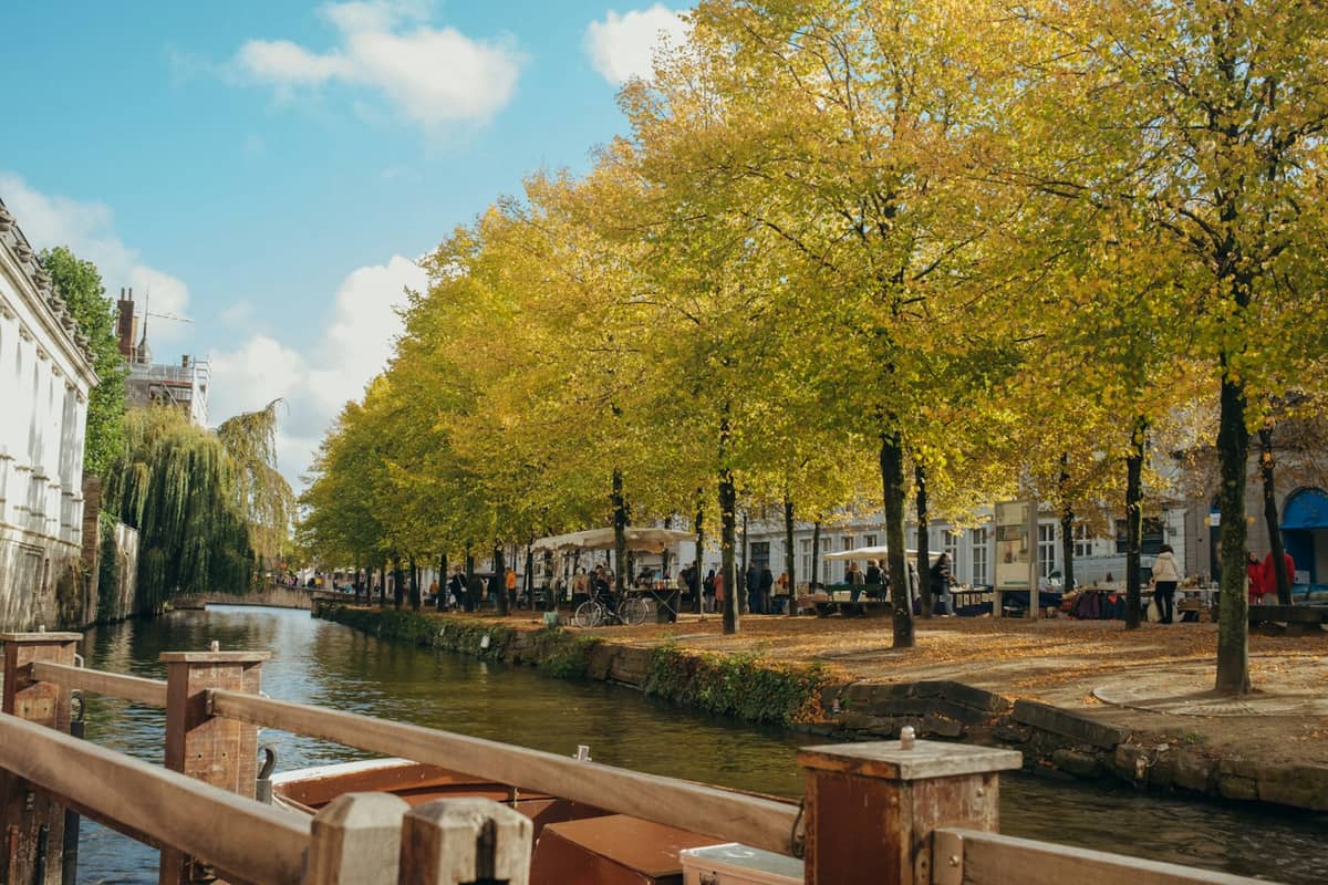 Bruges canal lined with trees showing early autumn yellow leaves, with people walking along the bank and blue sky overhead.