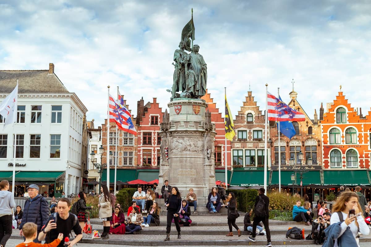 Crowds of people gathered around the Markt square and statue in Bruges on a bright summer day, with historic Flemish buildings in the background.