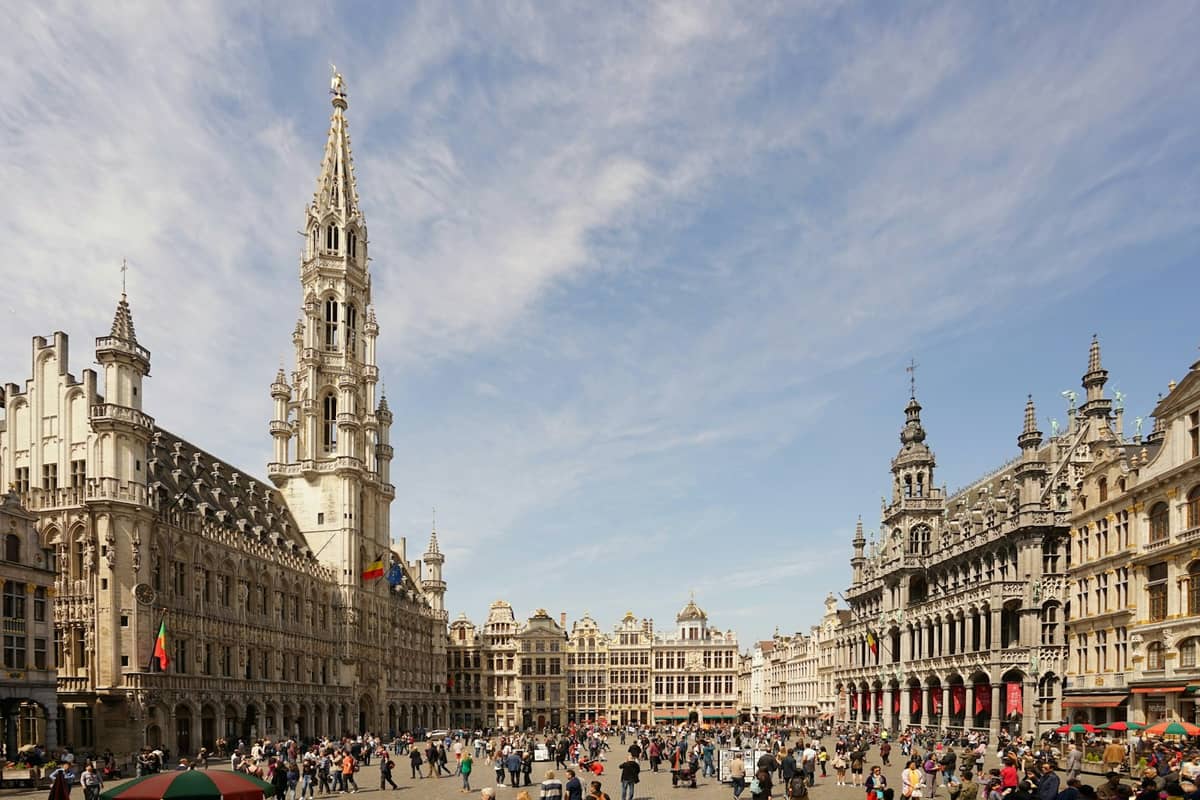 Vista ampla da Grand Place de Bruxelas em um dia ensolarado, com as guildas históricas, a torre da Câmara Municipal e muitas pessoas circulando pela praça.