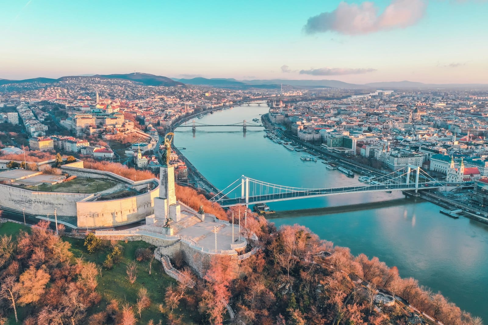 Aerial view of Budapest with the Danube River, bridges, and cityscape on a clear day, showcasing both Buda and Pest sides with vibrant colors and landmarks.