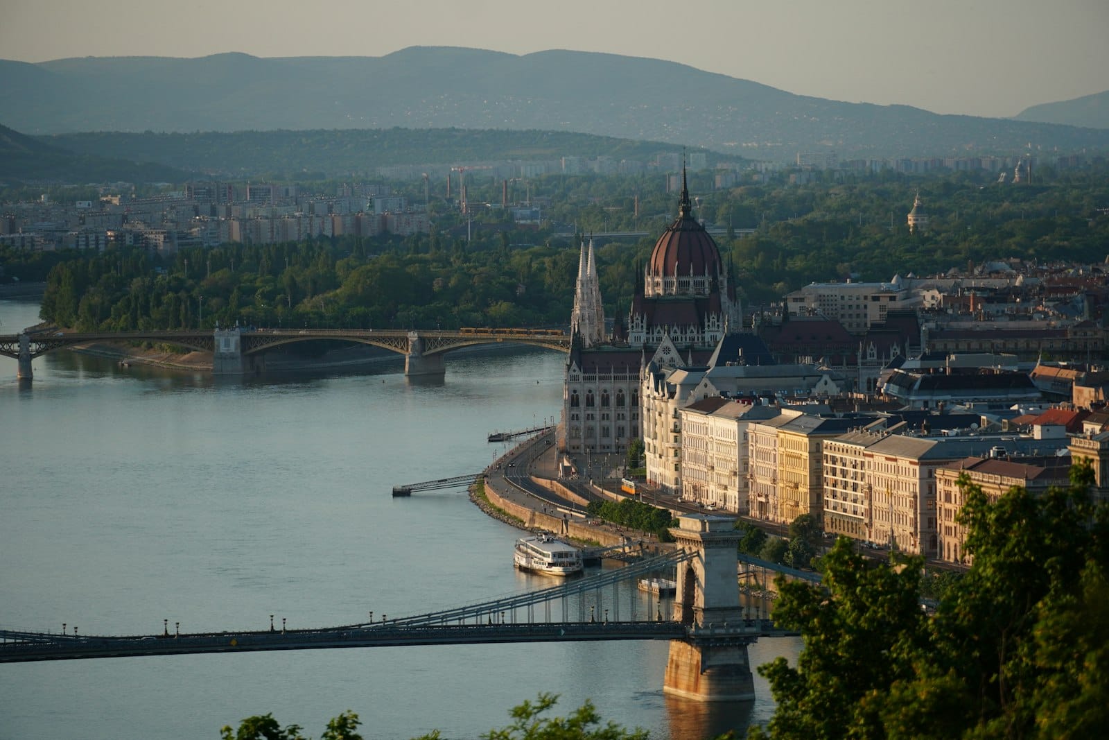 Panoramic view of Budapest with the Danube River, Parliament building, and bridges on a clear day, showing both Buda hills and Pest cityscape.
