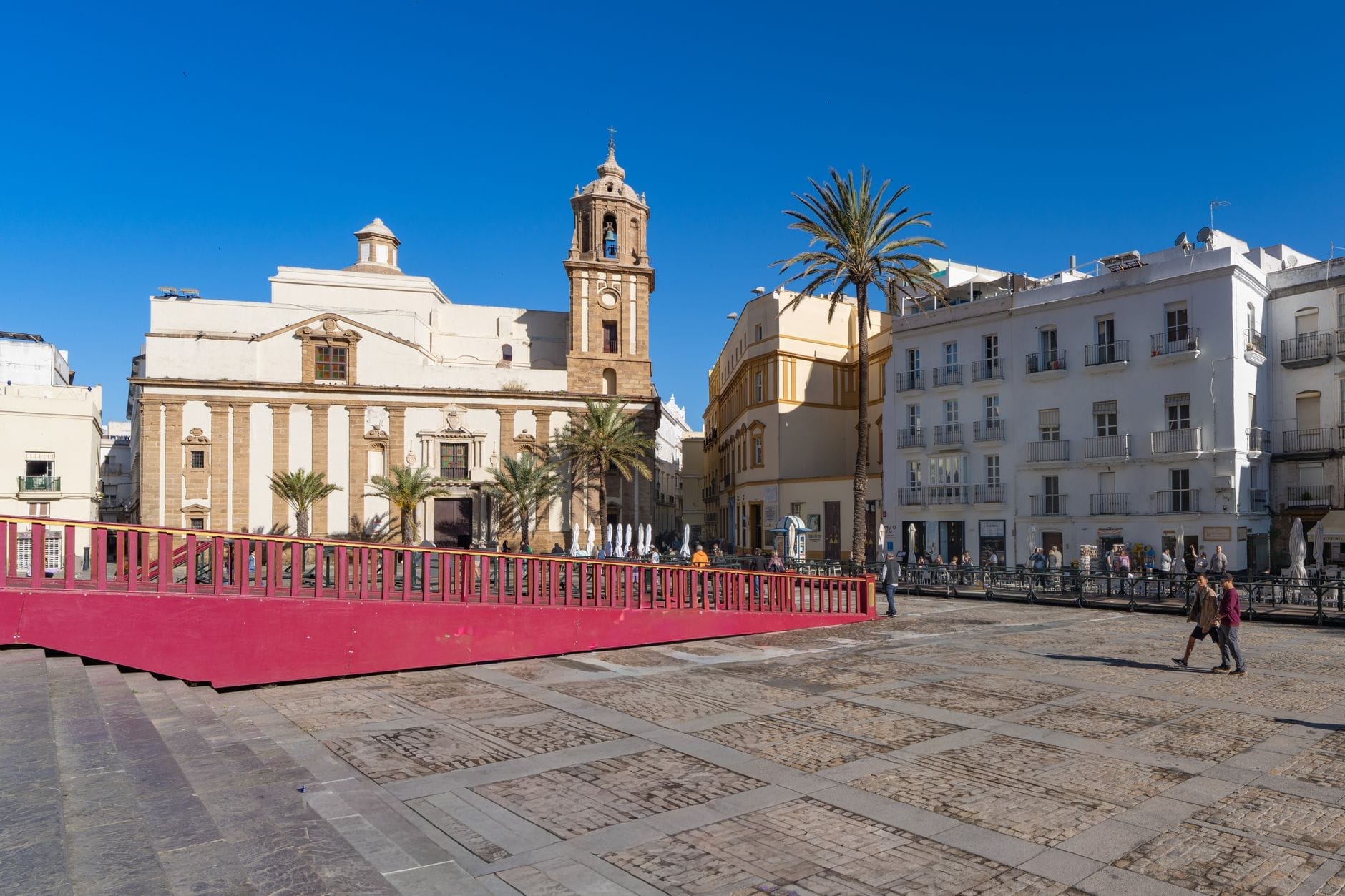 Praça aberta em Cádiz com edifícios históricos, uma igreja, uma palmeira e um céu azul brilhante de primavera, sugerindo clima primaveril agradável e facilidade para caminhar.