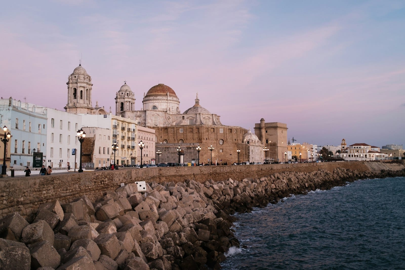 A orla marítima e a catedral de Cádiz, Espanha, ao entardecer com céus outonais em tons pastel e mar calmo ao longo do quebra-mar de pedra.