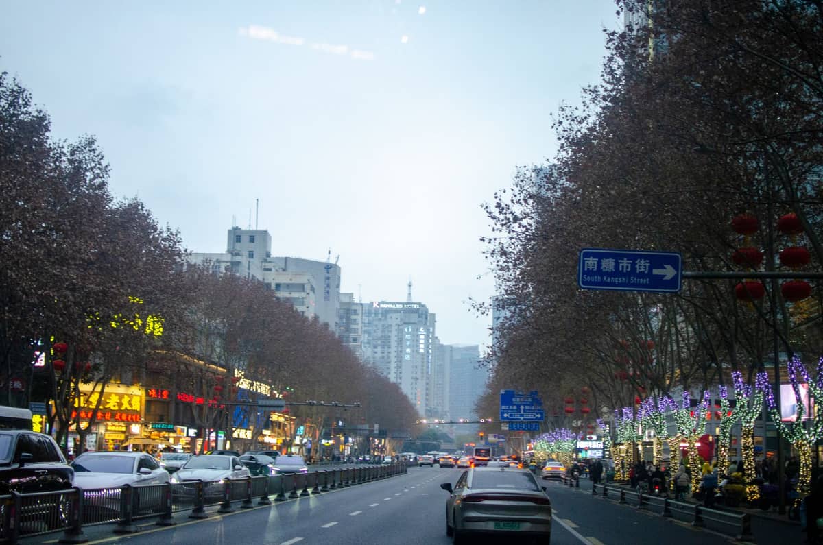 Une rue de Chengdu par une journée d'hiver grise et nuageuse avec des arbres dénudés, des voitures et des lumières festives longeant la route.