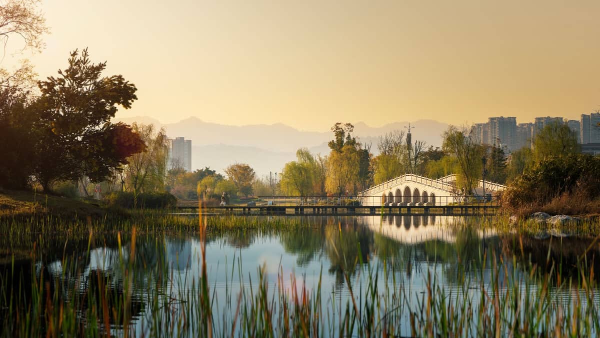Vue sereine d'un parc de Chengdu avec un pont en arc blanc sur un lac, des arbres, la skyline de la ville et des montagnes lointaines au lever ou au coucher du soleil.