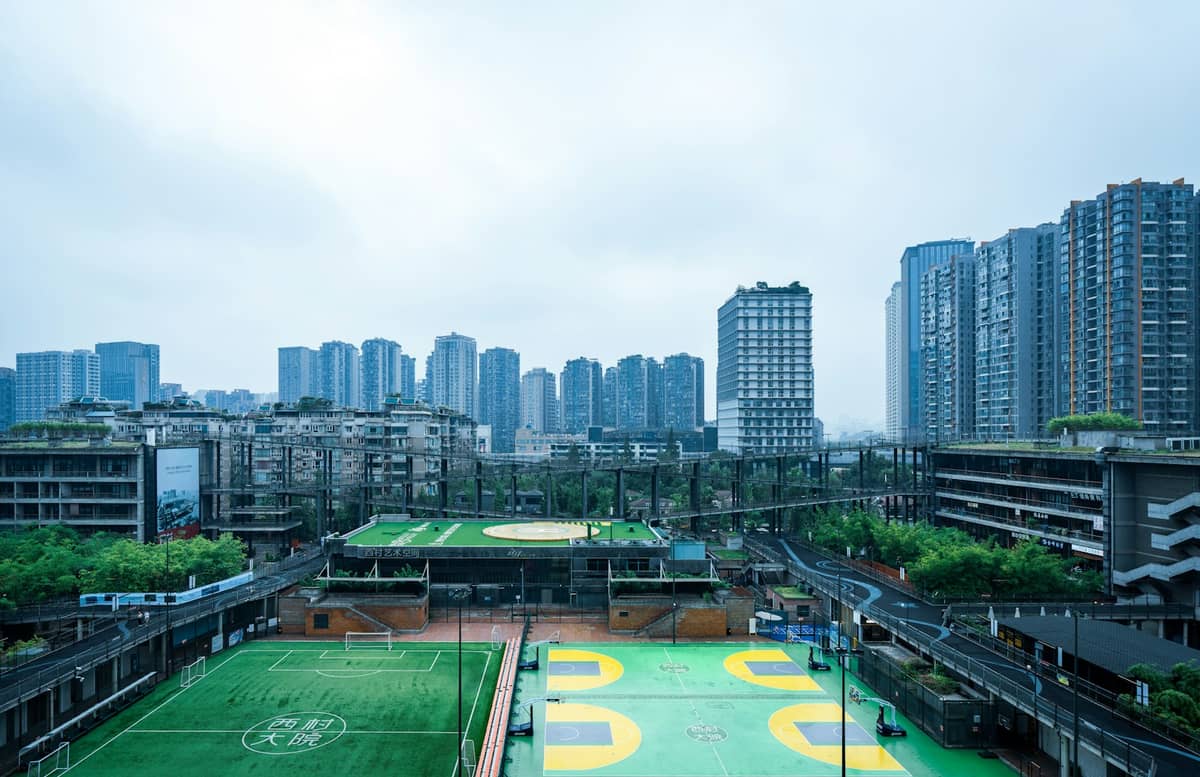 Large panorama urbain de Chengdu avec des immeubles de grande hauteur sous un ciel couvert, avec des espaces verts et des terrains de sport au premier plan.