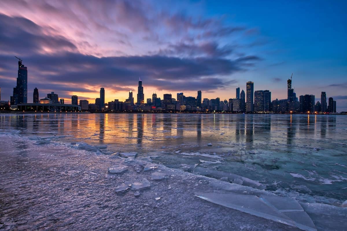 Zugefrorenes Seeufer mit Eis und dramatischen Wolken bei Sonnenaufgang, Skyline von Chicago im Hintergrund.