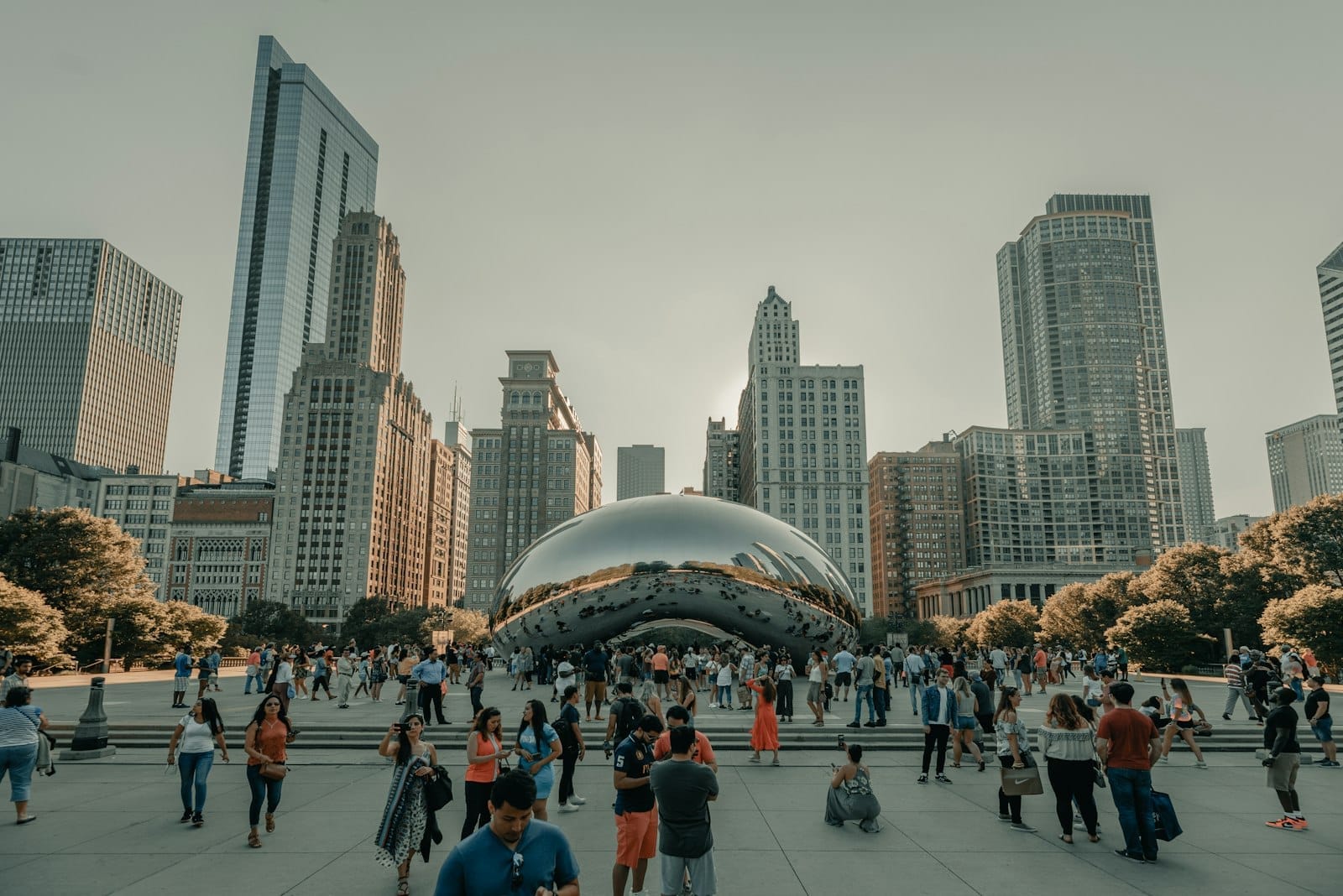 Uma multidão animada se reúne ao redor do icônico Cloud Gate no Millennium Park de Chicago em um dia ensolarado, com arranha-céus ao fundo.