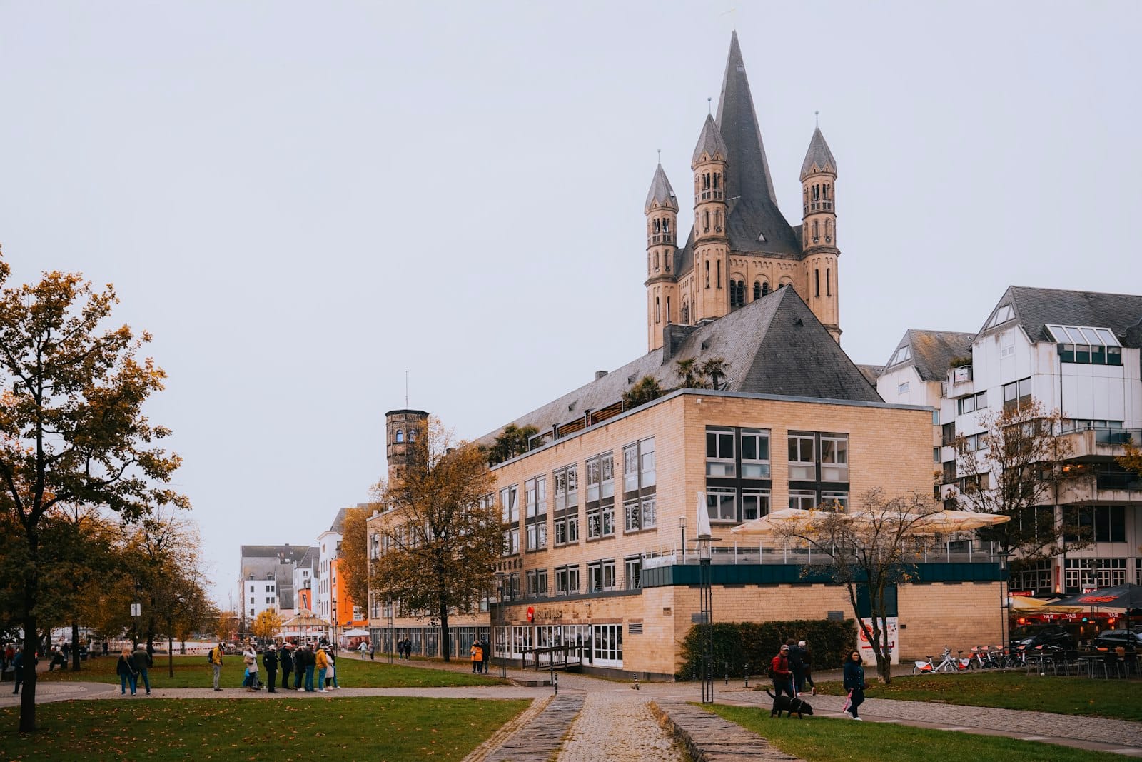 วิวทิวทัศน์ของโบสถ์ Groß St. Martin ในเมือง Cologne อยู่เบื้องหลังต้นไม้สีสันฤดูใบไม้ร่วงและผู้คนในสวนสาธารณะ สะท้อนบรรยากาศฤดูใบไม้ร่วงของเมือง