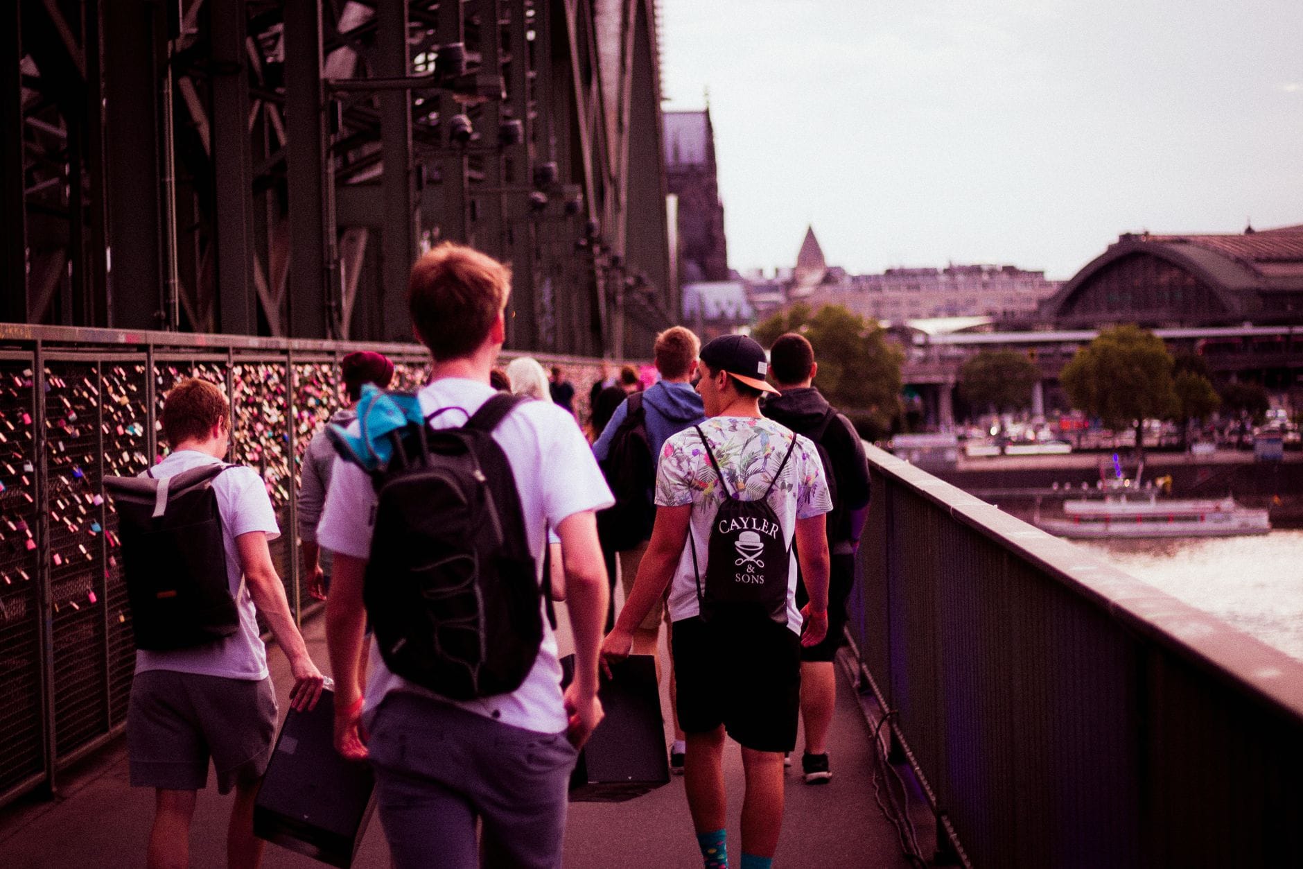 ผู้คนกำลังเดินบนสะพาน Hohenzollern ของเมือง Cologne ในวันที่แดดสดใส โดยมีตัวเมืองและแม่น้ำ Rhine มองเห็นได้เบื้องหลัง