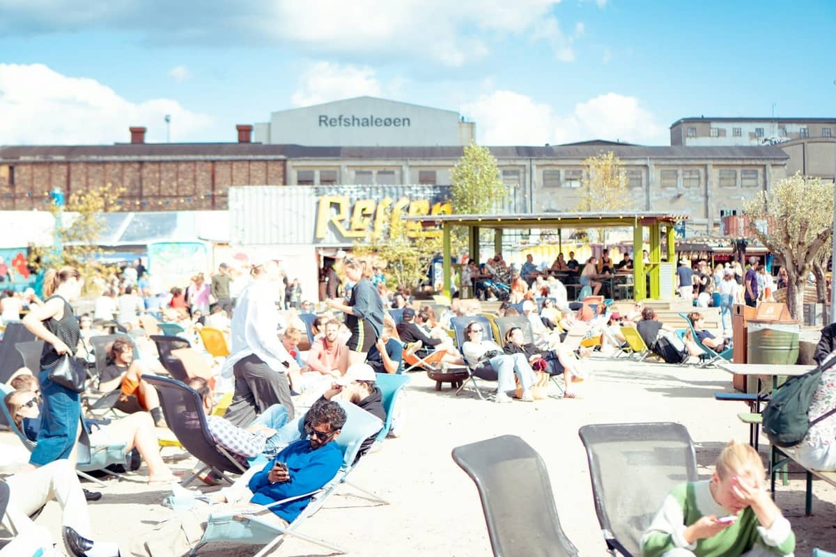 Foule se prélassant en plein air en été à Refshaleøen, Copenhague, avec des sièges festifs et des personnes profitant d'un événement animé sous un ciel bleu.