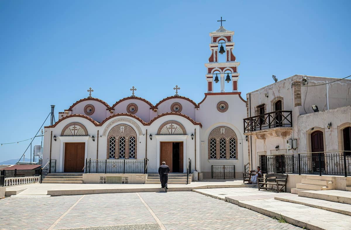 Weiße orthodoxe Kirche mit roten Kuppeln und Glockenturm auf einem sonnigen Dorfplatz auf Kreta, ein traditioneller Ort für lokale religiöse Feste.