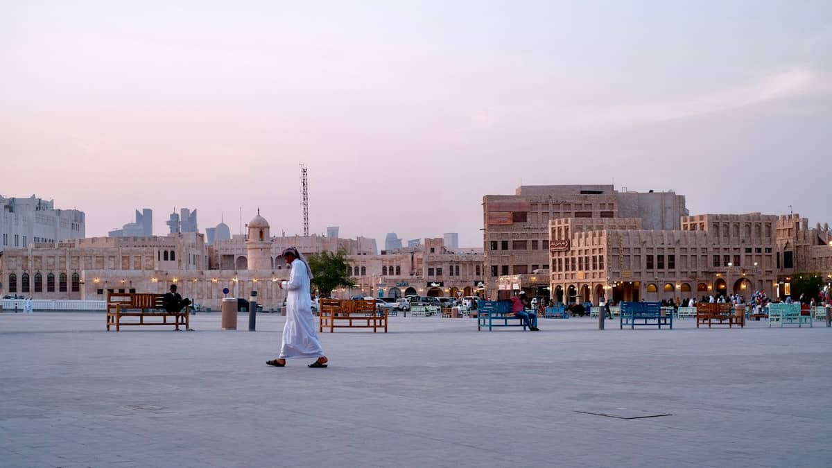 Ein Mann in traditioneller Kleidung überquert einen offenen Platz im Souq Waqif in Doha bei Sonnenuntergang, mit historischen Gebäuden im Hintergrund.