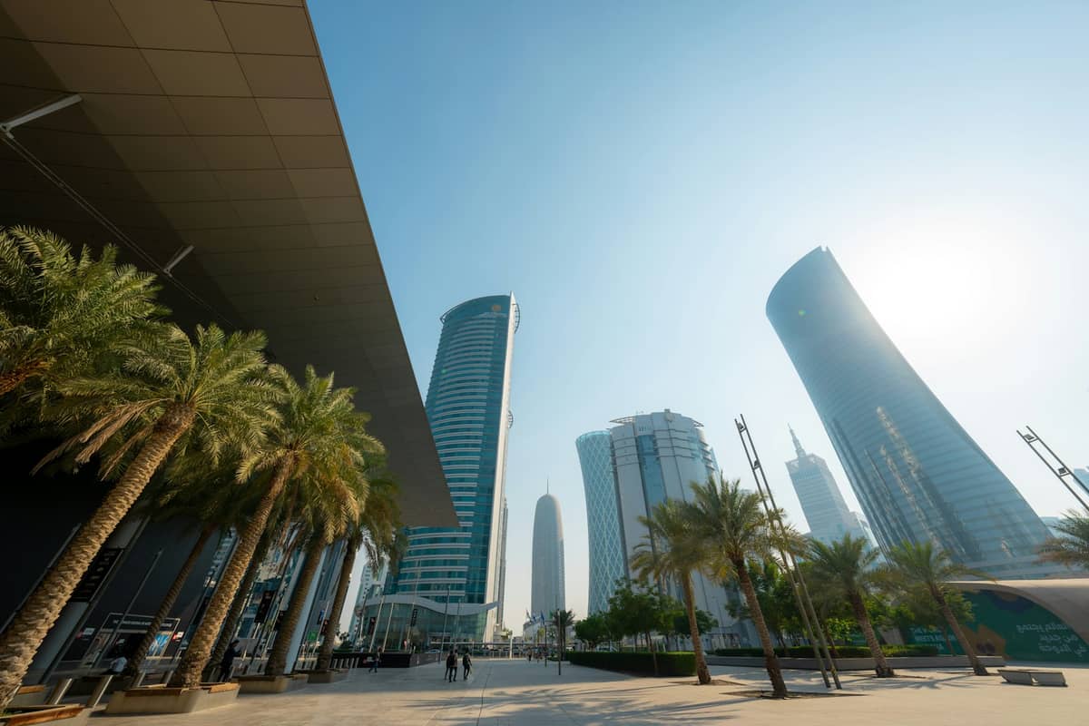 Moderne Wolkenkratzer in Doha mit Palmen unter einem hellen, dunstigen Sommerhimmel und einem sonnigen, leeren Platz