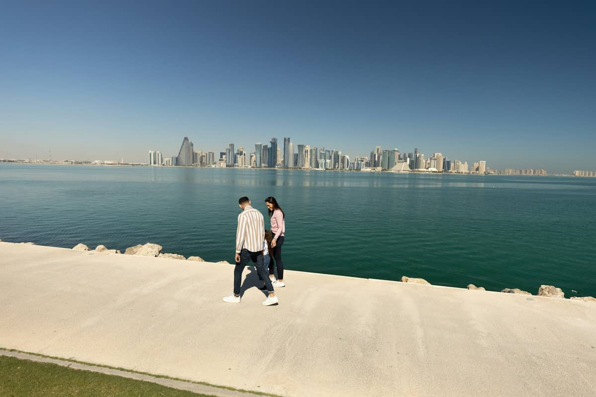 Zwei Personen spazieren entlang der Doha Corniche mit der Skyline der Stadt und dem Meer im Hintergrund unter klarem Himmel.