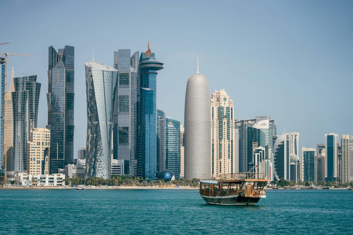 Weiter Blick auf die Skyline von Doha mit markanten modernen Wolkenkratzern und einem traditionellen hölzernen Dau-Boot auf der Bucht unter strahlend blauem Himmel.