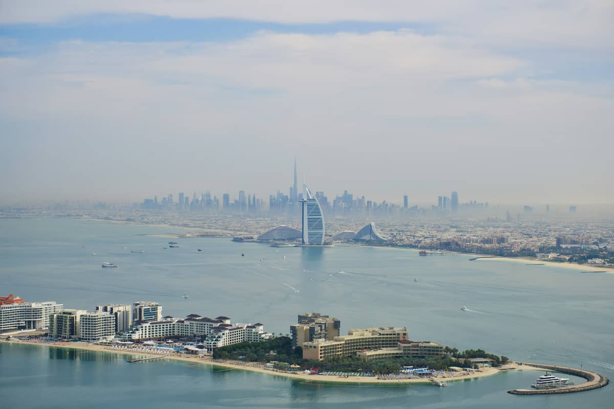 Luftaufnahme von Dubai mit Küstenlinie, Burj Al Arab, moderner Skyline und leichtem Dunst unter einem teilweise bewölkten Himmel.