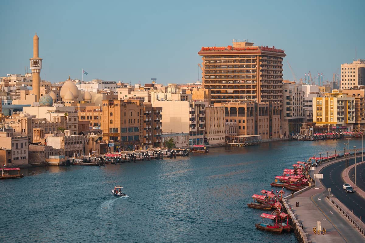 Weitläufiger Blick auf den Dubai Creek mit angedockten Abras, historischen Gebäuden, Minaretten und einer Uferpromenade an einem klaren, sonnigen Tag.