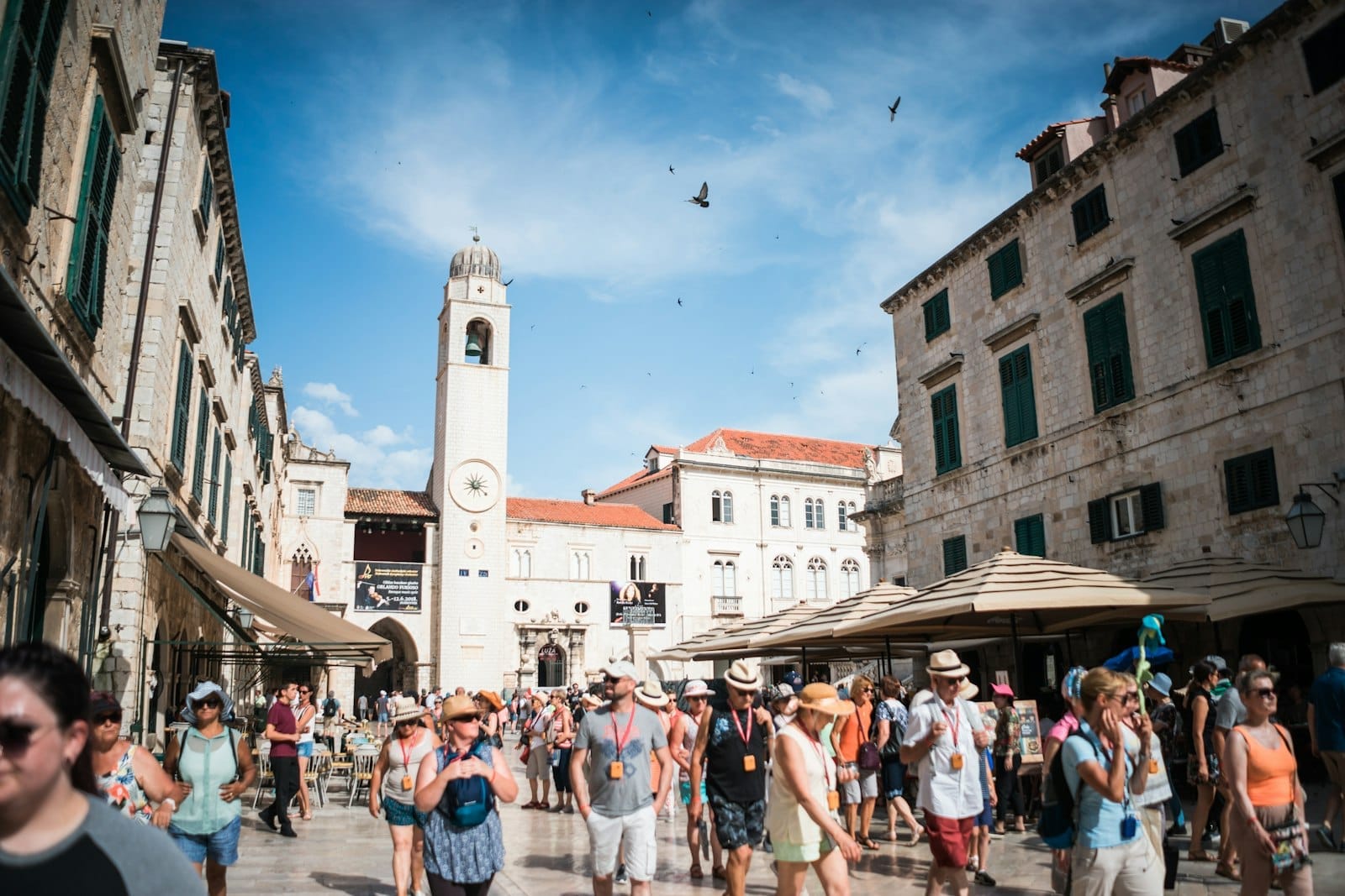 Rue piétonne animée dans la vieille ville de Dubrovnik avec des touristes se promenant sous un ciel lumineux, bâtiments en pierre et cafés visibles.