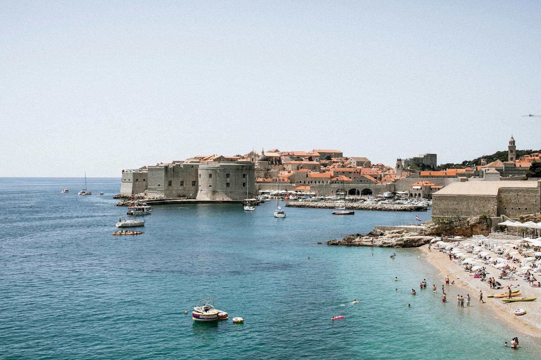 Vue sur les remparts de la vieille ville de Dubrovnik et une plage bondée avec des nageurs et des bateaux sous un ciel d'été éclatant.