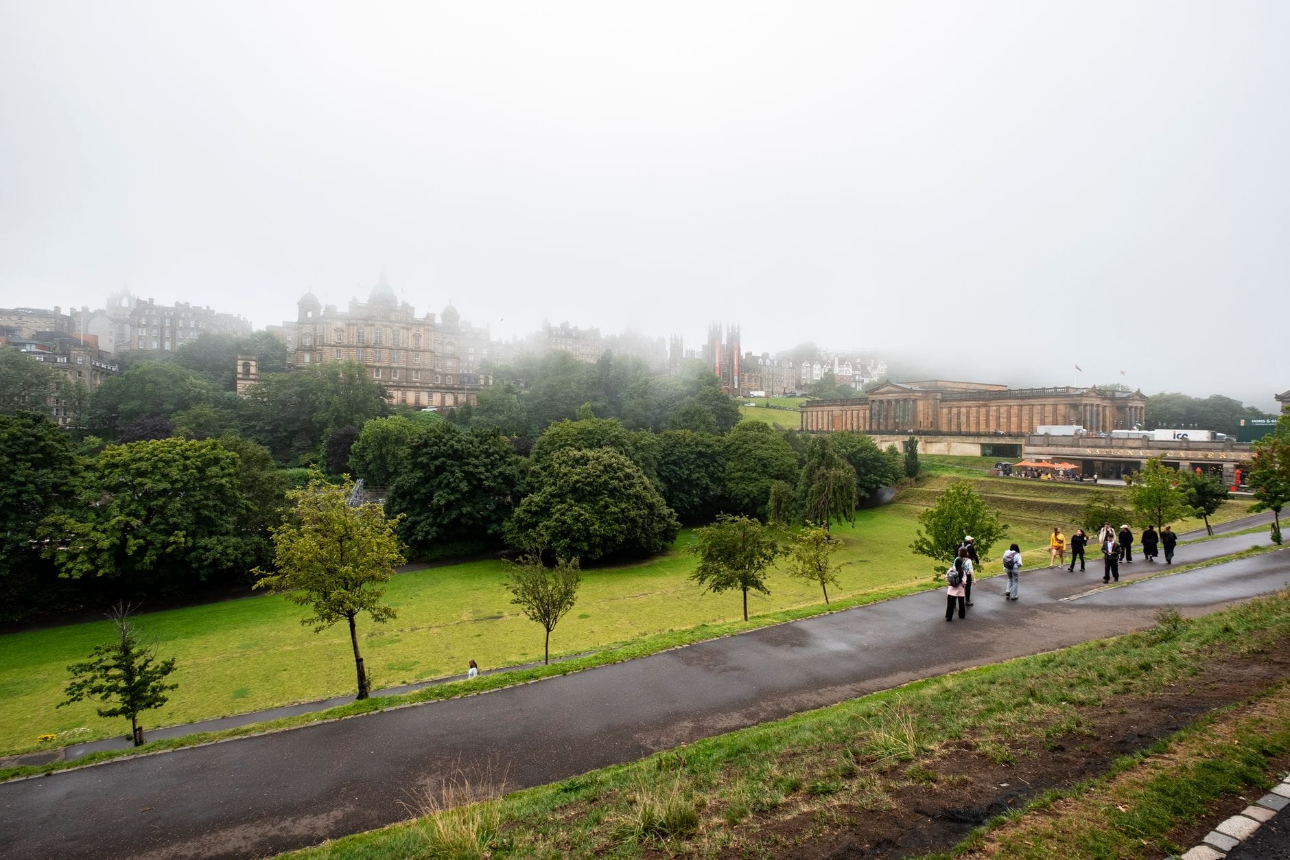 Vue de la ville d'Edinburgh avec des bâtiments historiques et des espaces verts, enveloppée de brume et de nuages gris bas, avec des sentiers humides et des piétons épars.