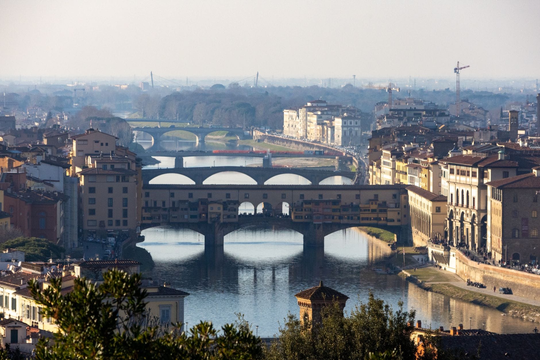 วิวพาโนรามาเหนือเมือง Florence แสดงให้เห็นสะพานหลายแห่ง แม่น้ำ Arno และทิวทัศน์เมืองที่ล้อมรอบด้วยเนินเขาในระยะไกลใต้ท้องฟ้าที่ขมุกขมัว