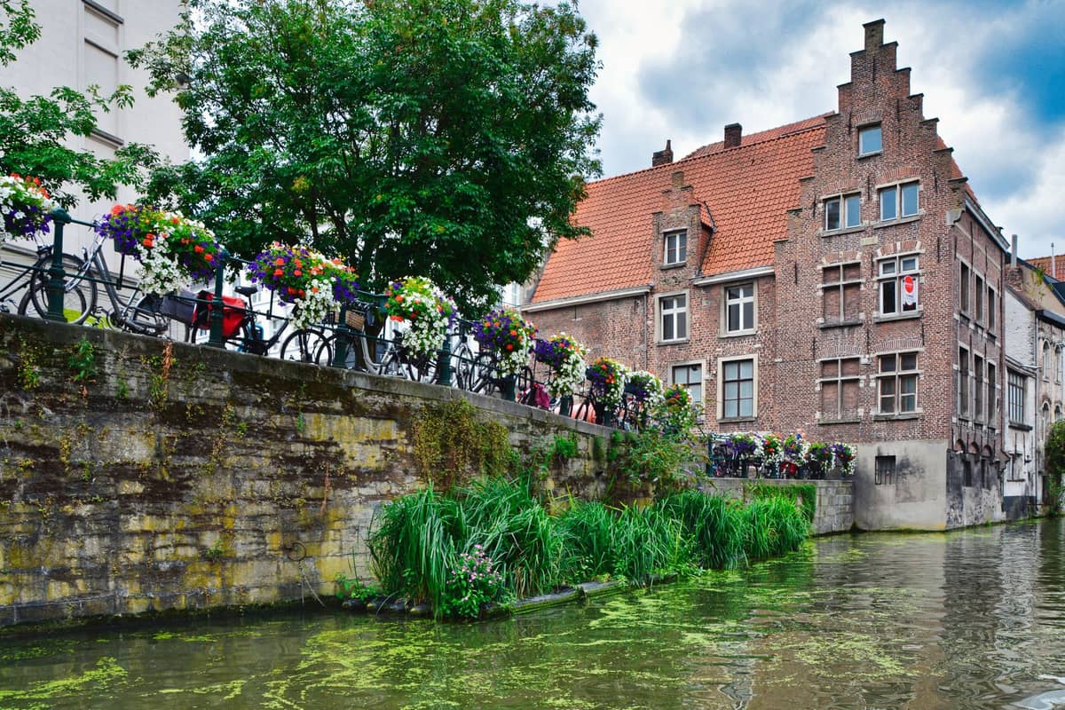Flower-lined canal with traditional Flemish buildings and greenery along the waterside in central Ghent on a bright day.