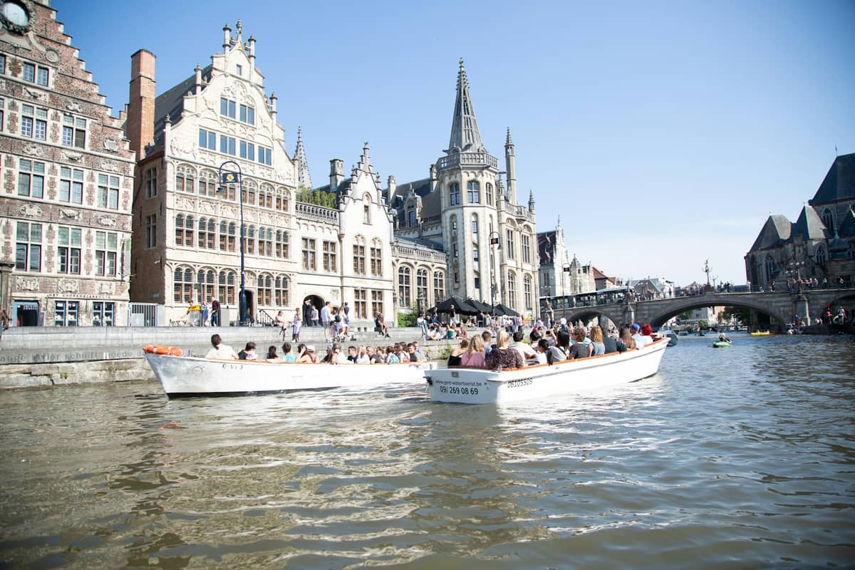 Sunny view of Ghent's Graslei waterfront with historic guildhalls, St. Michael’s Bridge, and tourist boats on the canal in spring.