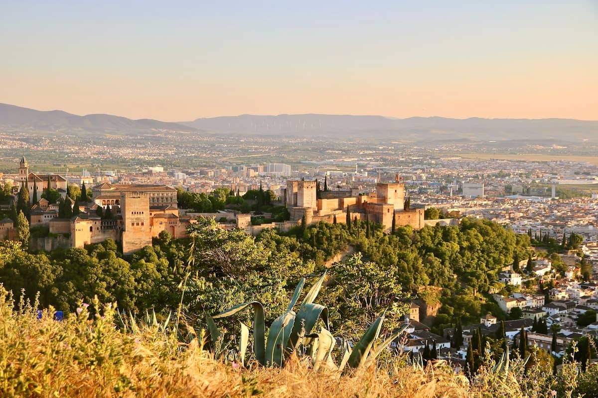 วิวยามอาทิตย์ทองเหนือ Granada พร้อม Alhambra และทิวทัศน์เมืองใต้ท้องฟ้าฤดูร้อนที่แจ่มใสและแดดจ้า มีพืชแห้งแล้งอยู่เบื้องหน้า