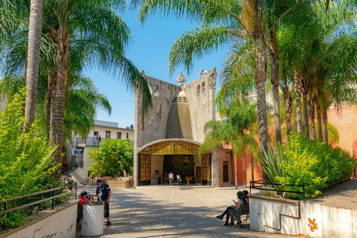 Ein sonnenbeschienener Platz in Guadalajara mit Palmen, Menschen, die sich auf Bänken entspannen, und einer markanten modernen Kirchenfassade im Hintergrund.