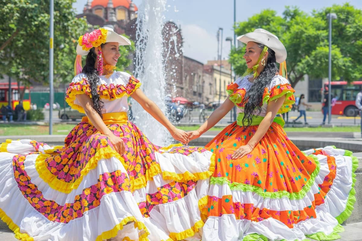 Zwei Frauen in bunten traditionellen mexikanischen Kleidern posieren und lächeln an einem Brunnen auf einem lebhaften Platz in Guadalajara, mit Bäumen und Kuppeln im Hintergrund.