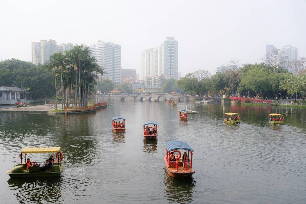 Grey, misty day with small boats on a lake in Guangzhou, city buildings and green trees in background.