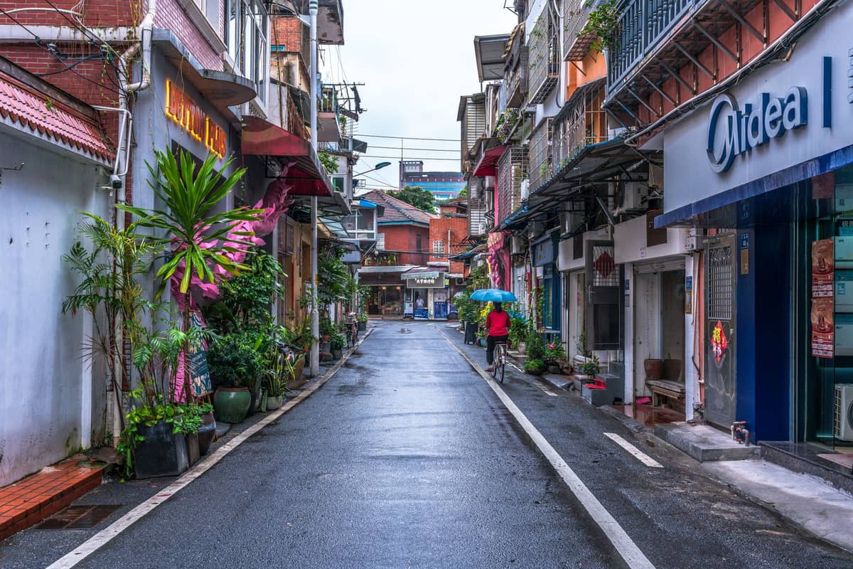 A wet, empty street in a historic Guangzhou neighborhood with a person holding an umbrella and humid summer atmosphere.
