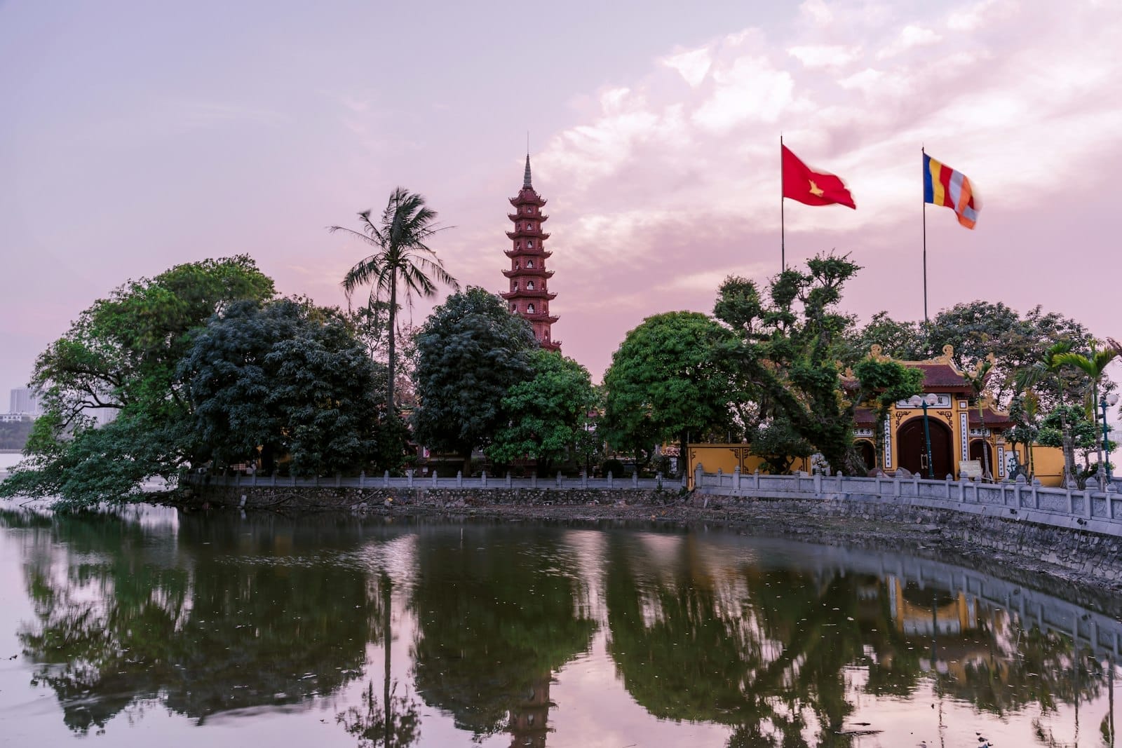 Tran Quoc Pagoda and its reflection in the lake at sunset in Hanoi, with vibrant flags and lush green trees under a colorful sky.