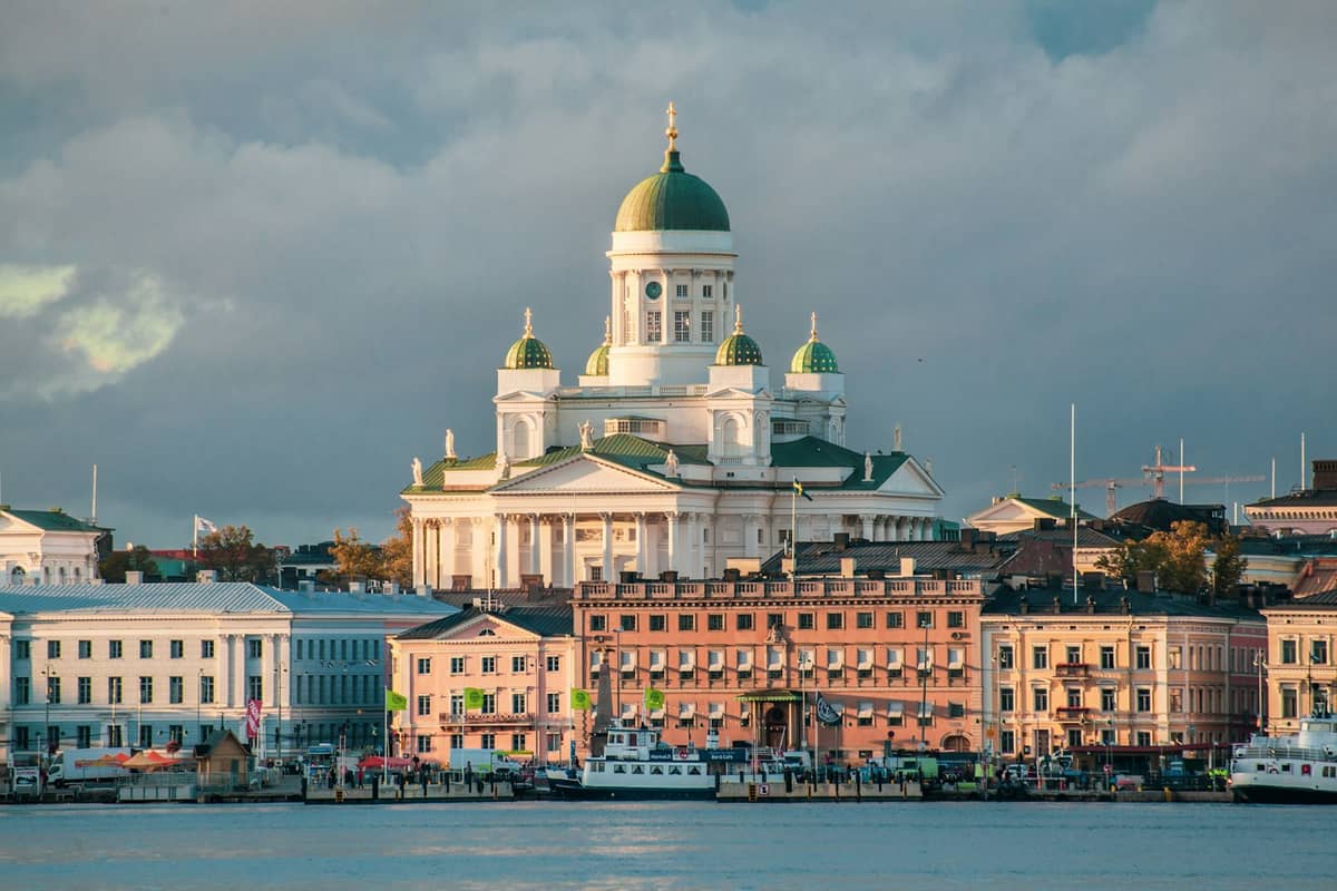 Vista do orla de Helsinque com edifícios históricos coloridos e a Catedral de Helsinque se destacando ao fundo sob um céu dramático.