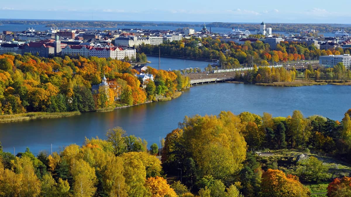 Vista panorâmica de Helsinki no outono mostrando árvores coloridas, edifícios da cidade, água e um céu claro e luminoso.
